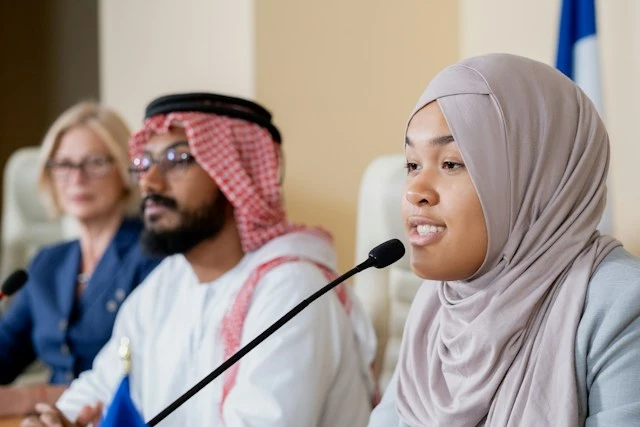 A woman wearing a hijab speaking into a microphone at an international panel discussion.